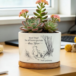 May include: A white ceramic cylindrical planter with a bamboo saucer containing three green succulents with small pink flowers. The planter features a black line drawing of Winnie the Pooh and Piglet with the text: Never forget The difference you make Mrs. Gloria Thank you, Brenda.