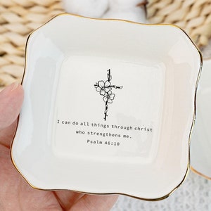 May include: Two white ceramic trinket dishes with gold trim. Each dish features a black and white cross with flowers and the text "I can do all things through christ who strengthens me. Psalm 46:10"