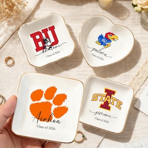 Pode incluir: Quatro pratos de cerâmica branca para bugigangas com bordas douradas. Um prato quadrado apresenta uma pegada laranja e o nome "Ainhoa" e "Class of 2026". Outro prato quadrado tem "BJ" e "khalani" com "Class of 2026". Um prato em forma de coração tem um logotipo de pássaro e "paloma" com "Class of 2026". O último prato tem "STATE" e "simone" com "Class of 2026".