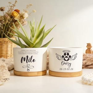 May include: Two white ceramic cylindrical planters with bamboo saucers. The left planter features the text "There is no greater earthly privilege than to have been loved by a dog," the name "Milo," and an infinity symbol with a paw print. The right planter displays a winged paw print with a halo, the name "Daisy," and the date "06.19-02.26."