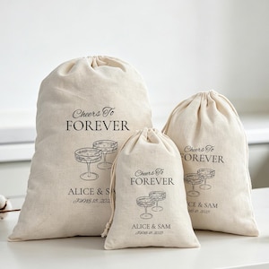 May include: Three off-white drawstring canvas bags of varying sizes, each printed with black text reading Cheers To FOREVER, ALICE & SAM, JUNE 18, 2025, and a line-art illustration of two stemmed cocktail glasses positioned side by side.