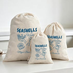 May include: Three off-white drawstring canvas bags of varying sizes, each featuring blue printed text that reads SEASHELLS & Wedding Bells and Miya's Bachelorette, accompanied by illustrations of a bucket of drinks, a seashell, and a bell.
