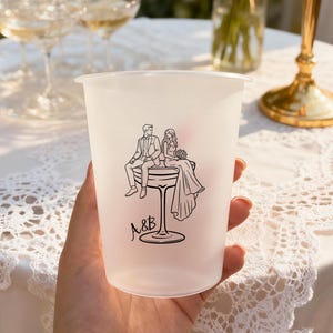 May include: A frosted plastic cup featuring a black and white illustration of a couple seated on a champagne glass. The initials "A&B" are below the illustration. The cup is held in a hand, with champagne glasses and a table in the background.