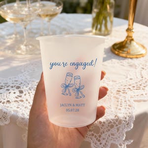 May include: A frosted plastic cup with the words "you're engaged!" in blue script. Below are two champagne glasses with bows, and the names "JACLYN & MATT" with the date "05.07.28" in blue. The cup is held in a hand.