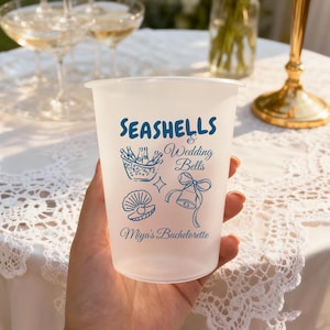 May include: A frosted plastic cup featuring blue printed graphics of a champagne bucket, a seashell with a pearl, and a bell with a bow. The text reads "SEASHELLS & Wedding Bells" and "Miya's Bachelorette" in a mix of block and script fonts.