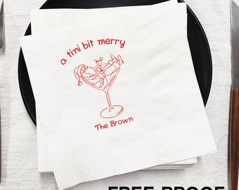 May include: White custom napkins with red text and a festive cocktail illustration. The text reads "a tini bit merry" and "The Brown". A black plate, fork, and knife are arranged around the napkins. The napkins are labeled "CUSTOM NAPKINS" and "FREE PROOF".