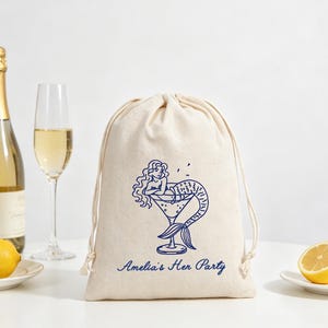 May include: A beige drawstring bag with a blue mermaid graphic in a martini glass. The text "Amelia's Her Party" is below the graphic. A bottle of champagne, a champagne flute, and lemons are in the background.