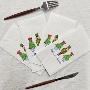 May include: Four white paper napkins with a Christmas theme. Each napkin features a watercolor illustration of a nutcracker holding a Christmas tree in a blue and white pot. The text "Mason Family" is printed on one of the napkins.