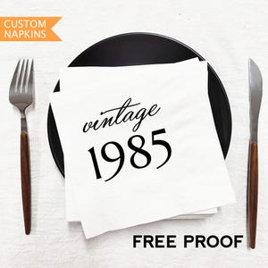 以下が含まれることがあります： 黒いプレートに重ねられた、黒い筆記体と太字で「vintage 1985」と書かれた白いカスタムナプキン。木製の柄が付いたフォークとナイフが両側にあります。「FREE PROOF」の文字が下にあります。