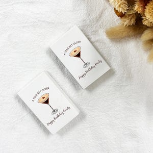 May include: Two white rectangular cards with a cocktail illustration and the text "A Tini Bit Older" and "Happy Birthday Emily". The cards are on a white textured surface, with dried flowers in the background.