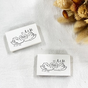 May include: Two white rectangular wedding favours with a black illustration of two swans facing each other, a heart, and the initials "A&M". The date "11.21.24" is printed below the swans. Dried flowers are in the background.