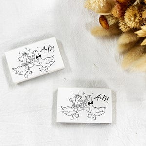 May include: Two rectangular white cards with a black line drawing of two geese toasting with wine glasses. The geese are wearing bow ties and floral headbands. The text "A+M" is written in black script on the right side of the card.