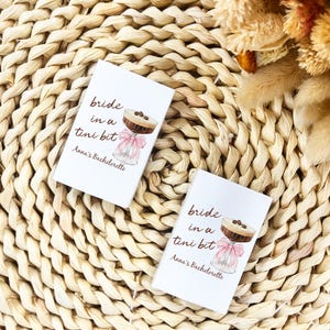 May include: Two white cards with the text "bride in a tini bit" and "Anna's Bachelorette" are displayed. Each card features a cocktail illustration with a pink bow. The cards are on a woven, beige placemat, with dried flowers in the background.