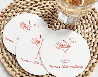 May include: Three white round coasters with a red cocktail illustration and the text "Emma's 30th Birthday". The coasters are on a woven placemat, with a glass of water and lemon slices in the background.