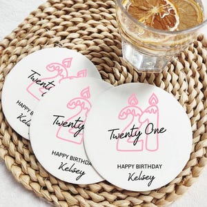 May include: White round coasters with pink "21" candle design and "Happy Birthday Kelsey" text. The coasters are on a woven placemat, with a glass of water and lemon slices in the background.