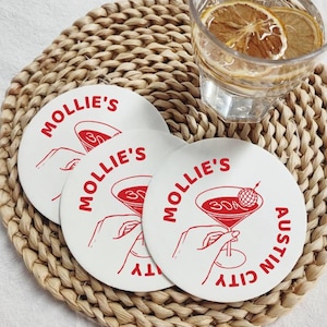 Puede incluir: Tres posavasos blancos con texto rojo y una ilustración de cóctel están dispuestos sobre un salvamanteles tejido. Los posavasos dicen "Mollie's Austin City" y presentan un diseño de copa de martini. Un vaso de agua con rodajas de limón está cerca.