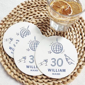 May include: Three white round paper coasters with blue illustrations of a disco ball, a martini glass, and a hand holding a bottle of champagne. The coasters have the text "William 06.24.2025" and the numbers "30" and "3" printed on them.