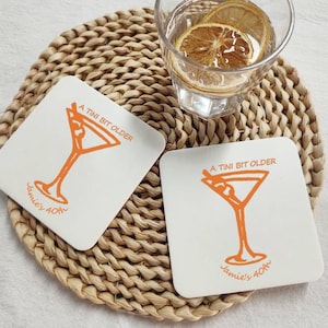 May include: Two white square coasters with orange text and a martini glass design. The text reads "A Tini Bit Older" and "Jamie's 40th".