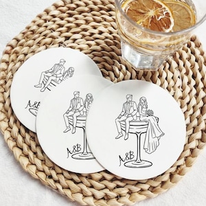 May include: White ceramic coasters with a black line drawing of a couple sitting on a champagne glass. The coasters have the initials "A&B" and are on a woven placemat. A glass of water with lemon slices is in the background.