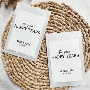 Könnte beinhalten: Zwei weiße Seidenpapier-Päckchen mit schwarzem Text, der "for your HAPPY TEARS" und "MIKE & LISA 26.06.2025" lautet. Die Päckchen liegen auf einer geflochtenen Strohmatte.