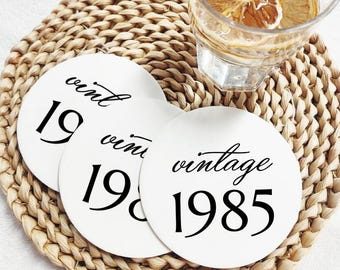 May include: Three white round coasters with the word "vintage" and the year "1985" printed in black. The coasters are on a woven placemat, with a glass of water and lemon slices in the background.
