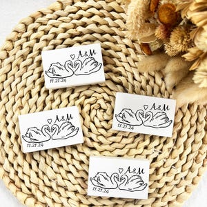 May include: Four white rectangular wedding favours with a black swan design, the initials "A&M", a heart, and the date "11.21.24". The favours are arranged on a woven, circular placemat with dried flowers in the background.