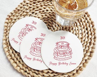 以下が含まれることがあります： 赤いケーキのデザインと「30！」と「Happy Birthday Emma」の文字が入った、白い丸いコースターが3つ。 コースターは織りのプレースマットの上にあり、背景には水とレモンスライスの入ったグラスがあります。