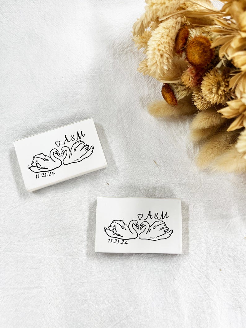May include: Two white rectangular wedding favours with a black illustration of two swans facing each other, a heart, and the initials "A&M". The date "11.21.24" is printed below the swans. Dried flowers are in the background.