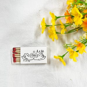 May include: A white matchbox featuring a black illustration of two swans, the initials "A&M", and the date "11.21.24". The match heads are red. Yellow and orange flowers are in the background.