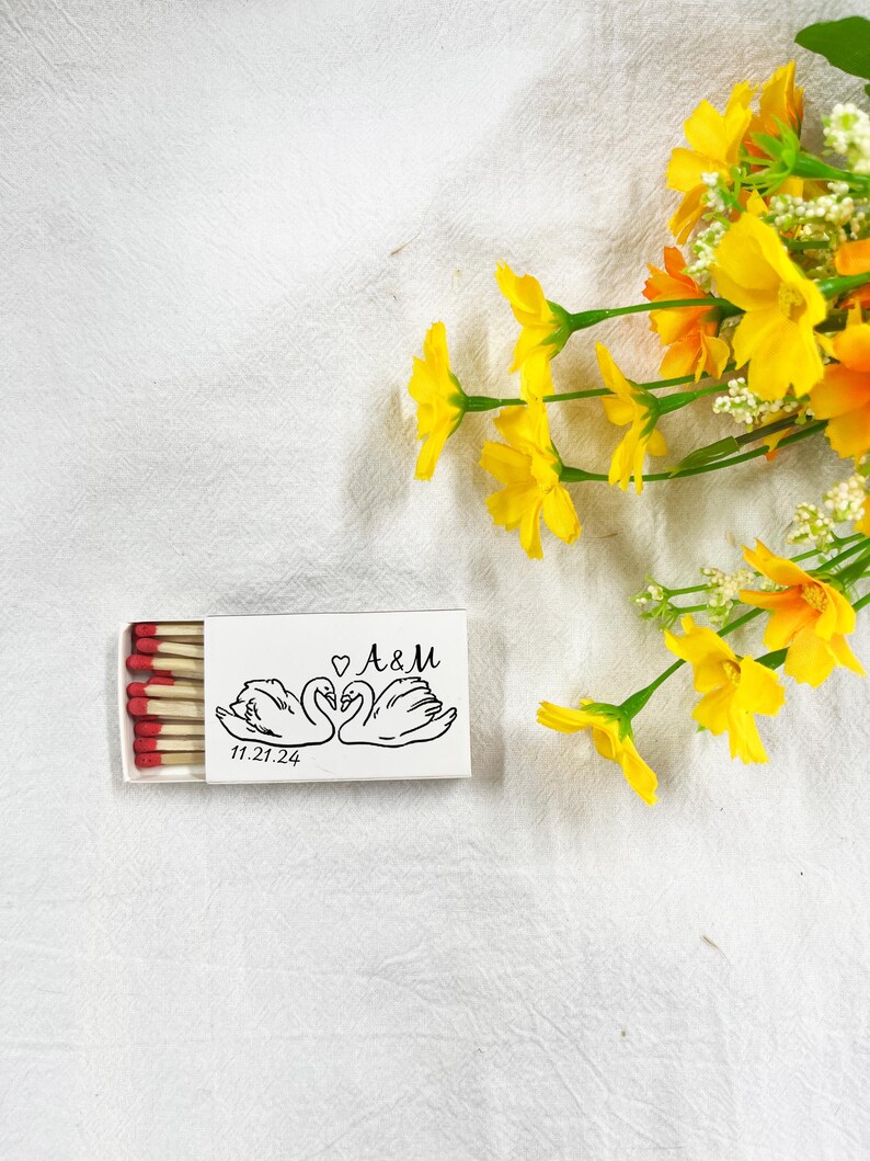 May include: A white matchbox featuring a black illustration of two swans, the initials "A&M", and the date "11.21.24". The match heads are red. Yellow and orange flowers are in the background.