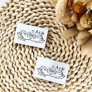 May include: Two white rectangular cards featuring a black swan illustration, the initials "A&M" with a heart, and the date "11.21.24". They rest on a woven, beige placemat, with dried flowers visible in the background.