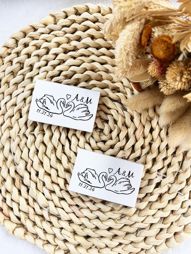 May include: Two white rectangular cards featuring a black swan illustration, the initials "A&M" with a heart, and the date "11.21.24". They rest on a woven, beige placemat, with dried flowers visible in the background.