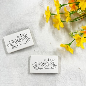 May include: Two white rectangular boxes featuring a black swan design, the initials "A&M" and the date "11.21.24". Yellow flowers are in the background. The swans are facing each other with a heart above them.