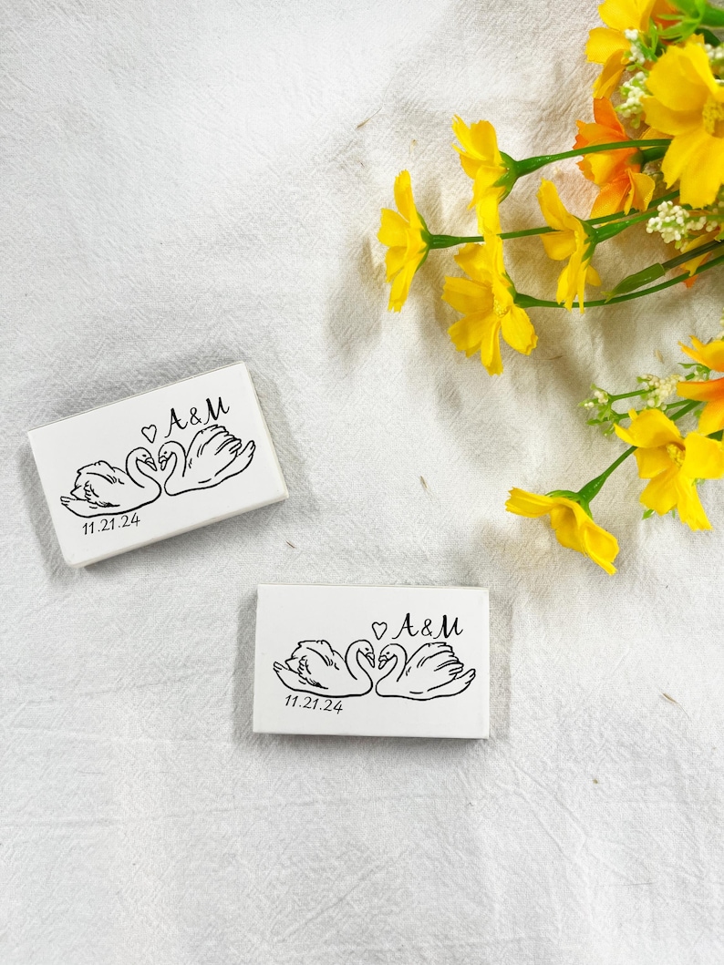 May include: Two white rectangular boxes featuring a black swan design, the initials "A&M" and the date "11.21.24". Yellow flowers are in the background. The swans are facing each other with a heart above them.