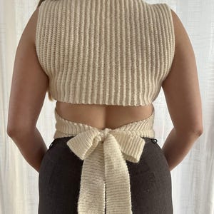 Peut inclure: Haut en tricot côtelé de couleur crème avec un détail de nœud dans le dos. Le haut sans manches a une encolure carrée et une conception enveloppante qui se noue en un nœud. Le tissu semble être une fibre naturelle et douce.