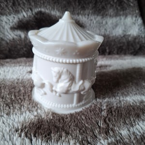 May include: White ceramic carousel-shaped jar with a lid. The jar features a detailed design of a carousel with horses and a canopy.