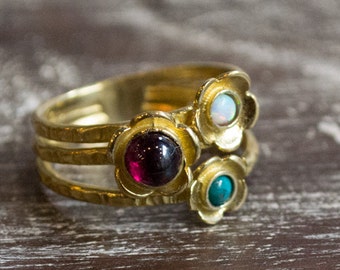 Könnte beinhalten: Ein goldfarbener Ring mit drei Bändern und blütenförmigen Fassungen. Die Fassungen halten einen dunkelroten Edelstein, einen weißen Opal und einen türkisfarbenen Stein. Der Ring liegt auf einer Holzoberfläche.