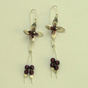 Pu&ograve; includere: Un paio di orecchini pendenti argentati. Ogni orecchino presenta un design a fiore a quattro petali con un centro rosso scuro. Sotto il fiore, una fila di piccole perline rosso scuro aggiunge eleganza. Chiusura a gancio.