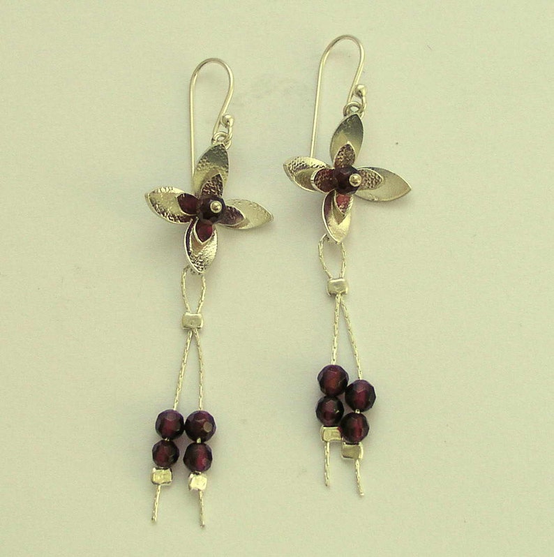 Pu&ograve; includere: Un paio di orecchini pendenti argentati. Ogni orecchino presenta un design a fiore a quattro petali con un centro rosso scuro. Sotto il fiore, una fila di piccole perline rosso scuro aggiunge eleganza. Chiusura a gancio.