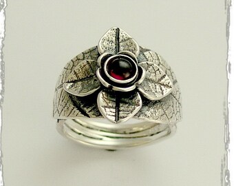 Puede incluir: Un anillo de plata con una piedra preciosa roja engastada en un diseño floral. El anillo está hecho de plata texturizada y tiene una banda que es ligeramente más ancha que el diseño floral.