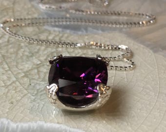 May include: A silver necklace with a large, square-cut, deep purple gemstone pendant. The pendant is set in a decorative silver setting with claw-like prongs. The chain is a delicate silver box chain. The necklace is displayed on a white surface.