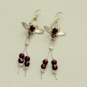 Pu&ograve; includere: Un paio di orecchini pendenti in argento. Ogni orecchino presenta un design floreale con un centro di perline rosso scuro e perline rosso scuro pendenti. Gli orecchini hanno una chiusura a gancio.