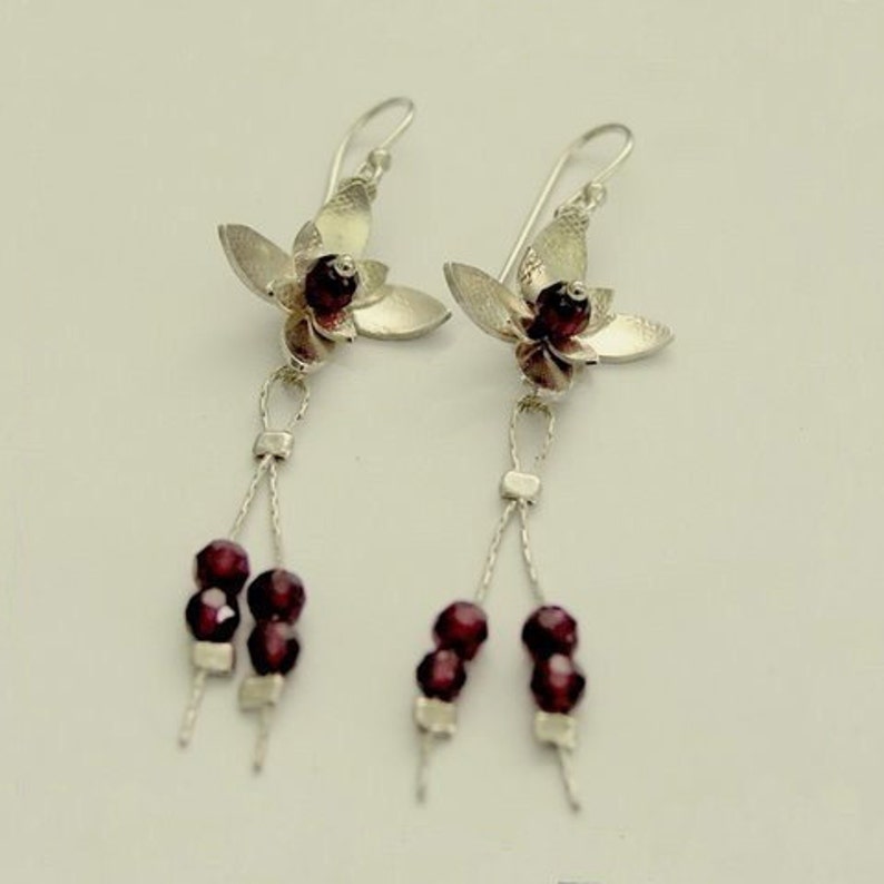 Pu&ograve; includere: Un paio di orecchini pendenti in argento. Ogni orecchino presenta un design floreale con un centro di perline rosso scuro e perline rosso scuro pendenti. Gli orecchini hanno una chiusura a gancio.