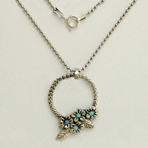 May include: Silver necklace with a circular pendant decorated with small flowers and leaves. The flowers have turquoise centres. The chain is made of small, round beads and has a clasp closure.