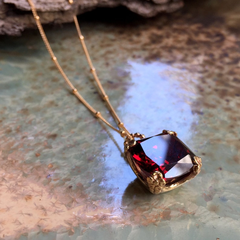 May include: A gold chain necklace with a large, square, red gemstone pendant. The pendant is set in a gold frame with intricate details.