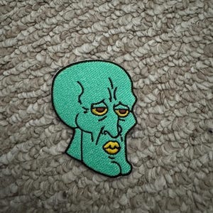 May include: Embroidered patch depicting a cartoon character with a green head, yellow eyes, and a pursed mouth. The patch has a black border and is set against a textured, neutral-toned background. The character's expression suggests a sense of melancholy.