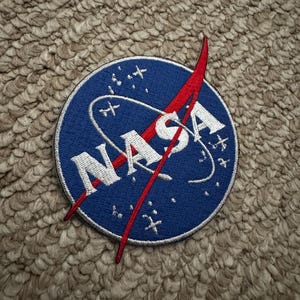 Peut inclure: Un &eacute;cusson circulaire avec le logo de la NASA. L'&eacute;cusson a un fond bleu avec le mot "NASA" en blanc et un graphique vectoriel rouge. Des &eacute;toiles blanches et une ligne blanche incurv&eacute;e sont &eacute;galement pr&eacute;sentes.