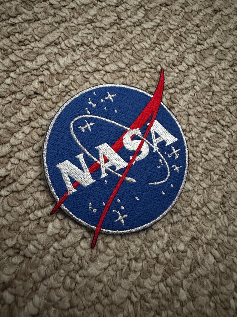 Peut inclure: Un &eacute;cusson circulaire avec le logo de la NASA. L'&eacute;cusson a un fond bleu avec le mot "NASA" en blanc et un graphique vectoriel rouge. Des &eacute;toiles blanches et une ligne blanche incurv&eacute;e sont &eacute;galement pr&eacute;sentes.
