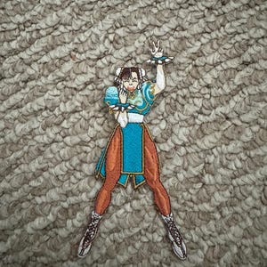 Może przedstawiać: Haftowana naszywka przedstawiająca postać żeńską w turkusowo-pomarańczowym stroju. Postać jest w dynamicznej pozie, z uniesioną ręką. Naszywka ma szczeg&oacute;łowe szwy i białą obw&oacute;dkę. Idealna do odzieży.