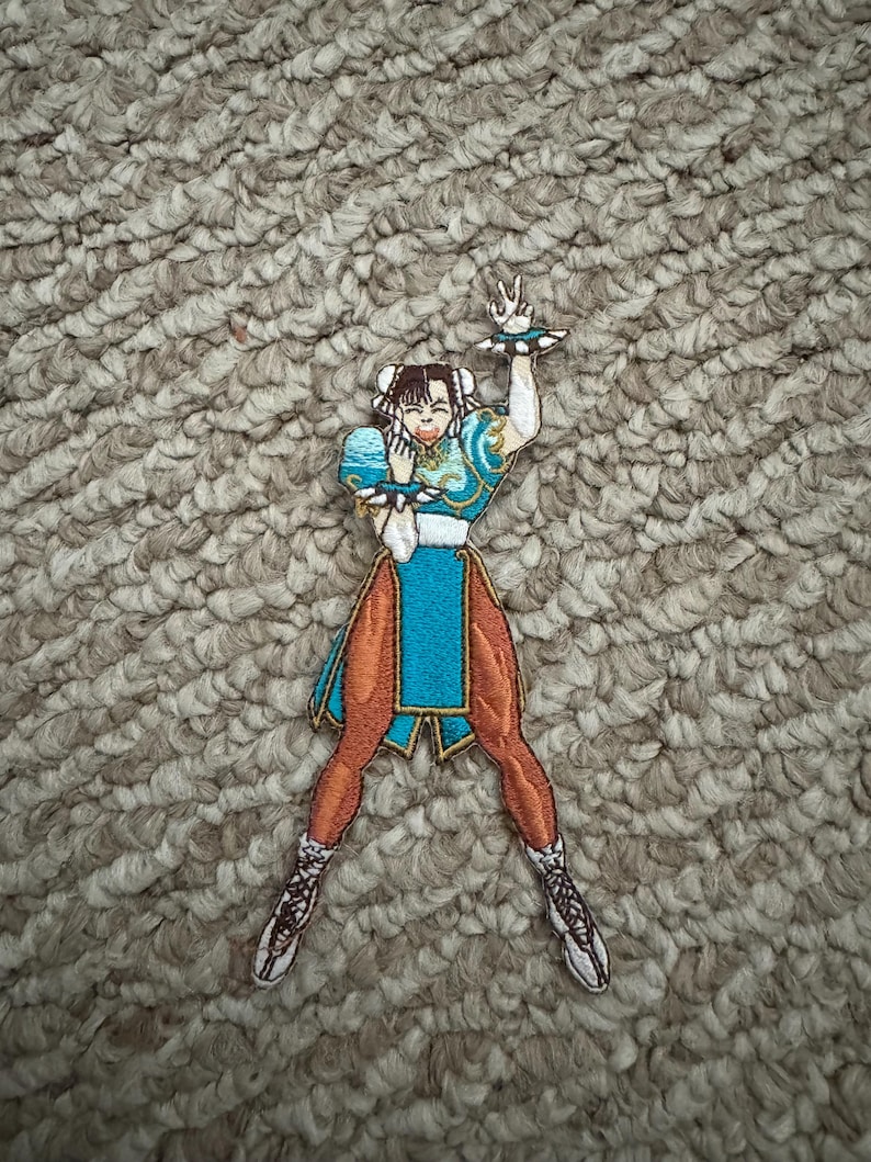 Może przedstawiać: Haftowana naszywka przedstawiająca postać żeńską w turkusowo-pomarańczowym stroju. Postać jest w dynamicznej pozie, z uniesioną ręką. Naszywka ma szczeg&oacute;łowe szwy i białą obw&oacute;dkę. Idealna do odzieży.